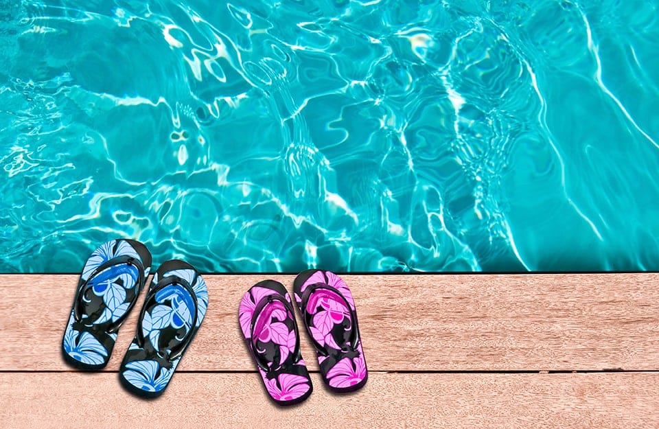 Vista dall'alto di due paia di infradito di colori diversi (azzurre e magenta) lasciate sulle assi in legno del decking a bordo piscina