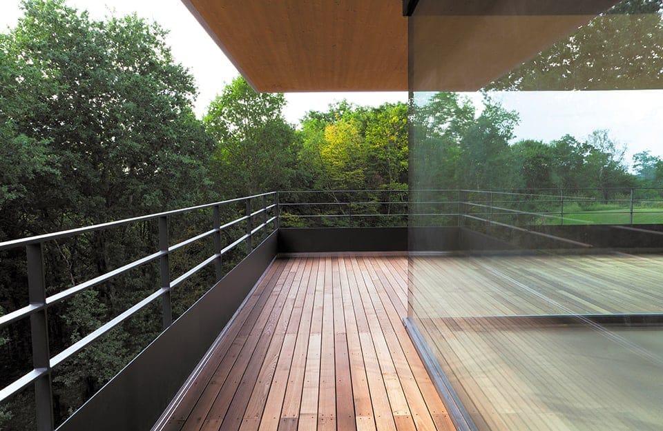 Terrazza esterna con pavimento in decking, ringhiere in metallo, e grande parete vetrata che separa l'interno dall'esterno, immerso nel verde degli alberi