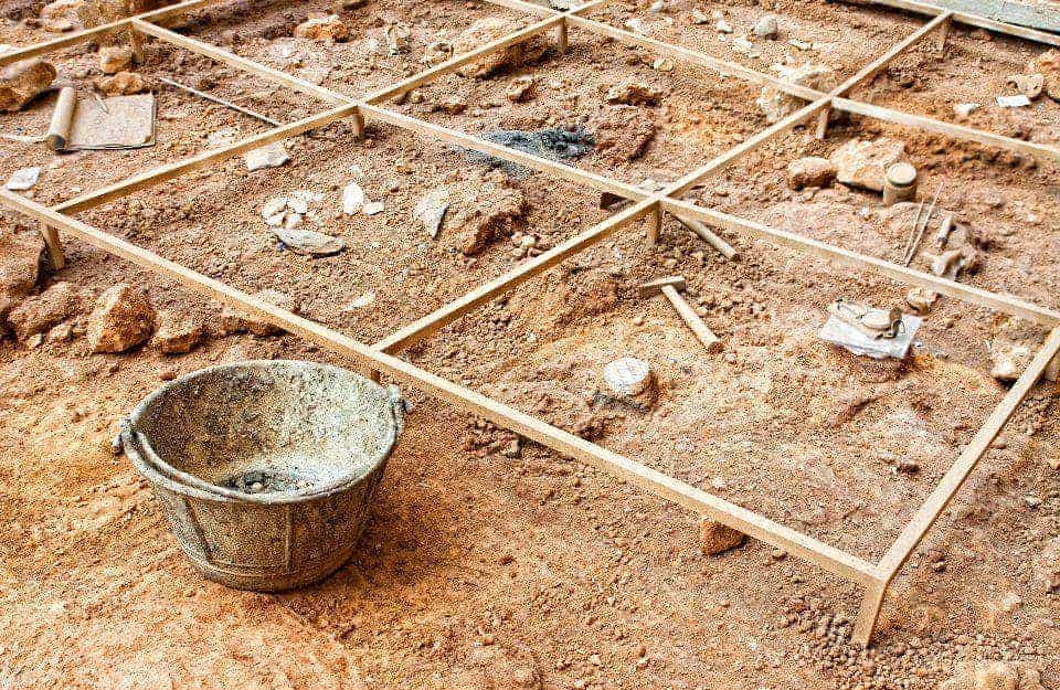 Parte di uno scavo archeologico, con una griglia in legno a suddividere in quadrati il terreno da scavare. Sono presenti anche secchi, martelli e picconi, oltre a piccoli reperti
