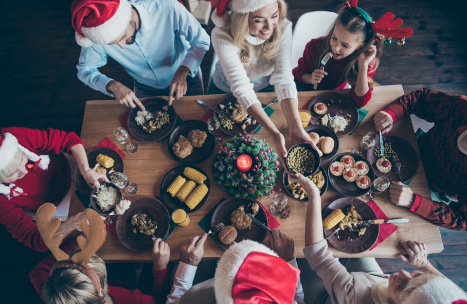 Vista dall'alto di una tavola imbandita per il Natale, con molte persone, grandi e bambini, che stanno pranzando con gioia indossando il cappello di Babbo Natale