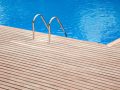 Una piscina con acqua blu e limpida. Sul bordo della piscina c'è un decking in teak. Al centro della piattaforma, c'è una scala in metallo con due corrimano curvi che scendono nell'acqua