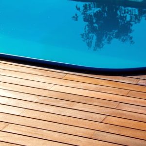 Sezione di una piscina con acqua limpida e azzurra e una pavimentazione in legno di teak intorno. Il bordo della piscina è curvo e l'acqua riflette alcuni alberi o piante. Il decking in teak è composto da assi orizzontali
