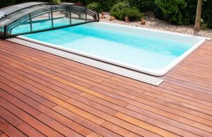 Una piscina rettangolare con acqua limpida e azzurra, circondata da un'ampia pavimentazione in legno di teak. La piscina ha una copertura trasparente parzialmente aperta, che può essere utilizzata per proteggere l'acqua e mantenere la temperatura. Sullo sfondo, ci sono piante verdi e un'area con ghiaia e decorazioni da giardino