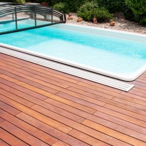 Una piscina rettangolare con acqua limpida e azzurra, circondata da un'ampia pavimentazione in legno di teak. La piscina ha una copertura trasparente parzialmente aperta, che può essere utilizzata per proteggere l'acqua e mantenere la temperatura. Sullo sfondo, ci sono piante verdi e un'area con ghiaia e decorazioni da giardino