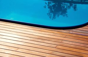 Sezione di una piscina con acqua limpida e azzurra e una pavimentazione in legno di teak intorno. Il bordo della piscina è curvo e l'acqua riflette alcuni alberi o piante. Il decking in teak è composto da assi orizzontali