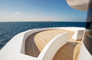 Il ponte di uno yacht o una barca moderna. Il ponte è fatto di tavole di lteak e ha un design elegante e curvo con ringhiere bianche. Lo yacht è in mare aperto, con una vasta distesa di acqua blu e una linea di costa distante visibile all'orizzonte. Il cielo è sereno con alcune nuvole sparse