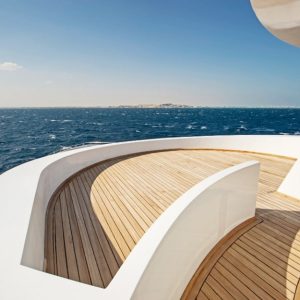 Il ponte di uno yacht o una barca moderna. Il ponte è fatto di tavole di lteak e ha un design elegante e curvo con ringhiere bianche. Lo yacht è in mare aperto, con una vasta distesa di acqua blu e una linea di costa distante visibile all'orizzonte. Il cielo è sereno con alcune nuvole sparse