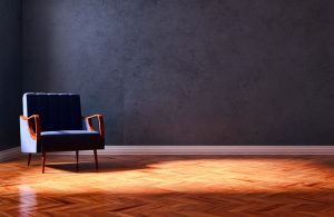 Poltrona in stile modernista con braccioli e gambe in legno naturale illuminata in maniera drammatica in una stanza vuota con parquet e pareti grigio scuro
