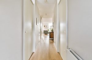 Un lungo e luminoso ma stretto corridoio di un grande appartamento, con porte da entrambi i lati, pareti chiare, pavimenti a parquet e salotto che si intravede in fondo