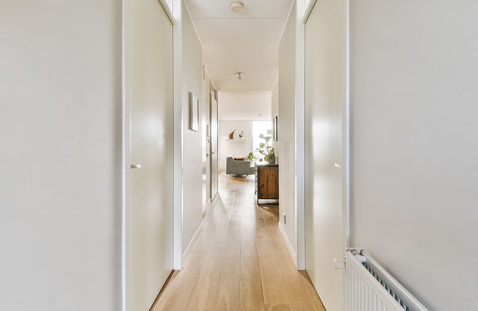 Un lungo e luminoso ma stretto corridoio di un grande appartamento, con porte da entrambi i lati, pareti chiare, pavimenti a parquet e salotto che si intravede in fondo
