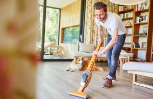 Un uomo pulisce un pavimento in parquet di un luminoso salotto con un'aspirapolvere. Il suo cane lo osserva