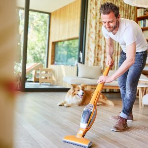 Un uomo pulisce un pavimento in parquet di un luminoso salotto con un'aspirapolvere. Il suo cane lo osserva