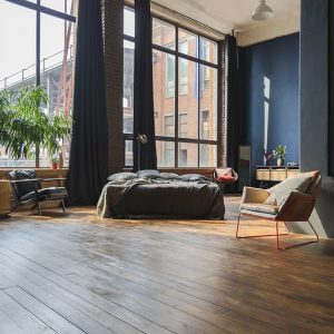 Grande open space con letto e poltrona. La luce entra da due grandi finestre che arrivano fino a terra. Il pavimento è un parquet rustico