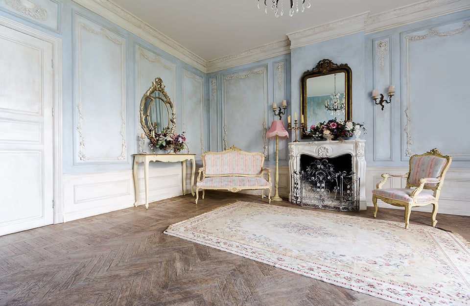 Ambiente shabby chic con arredi d'epoca riccamente decorati, pareti bianco e azzurro pastello, un grande tappeto davanti al caminetto e parquet rustico scuro