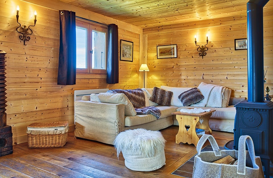 Salotto rustico da baita. Pavimento e pareti sono interamente in legno, le luci sono calde, ci sono una grande stufa blu in ghisa, un sofà d'angolo e un tavolino da caffè pure in stile rustico
