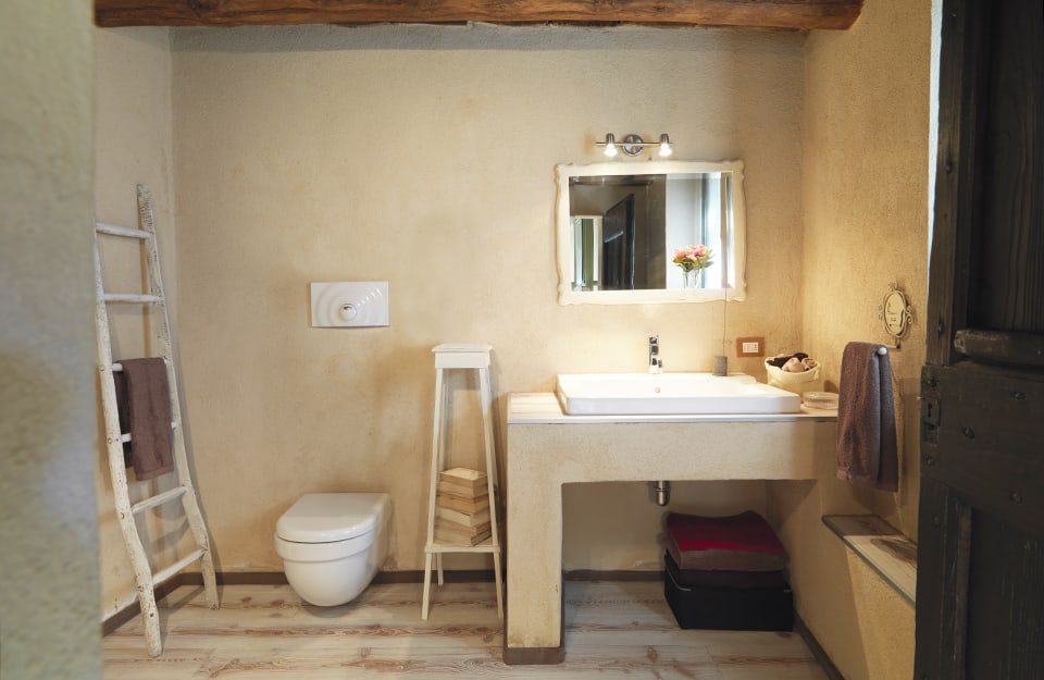 Bagno con elementi naturali: pietra, legno, metallo, travi a vista. Una vecchia scala dipinta di bianco appoggiata alla parete funge da porta asciugamani