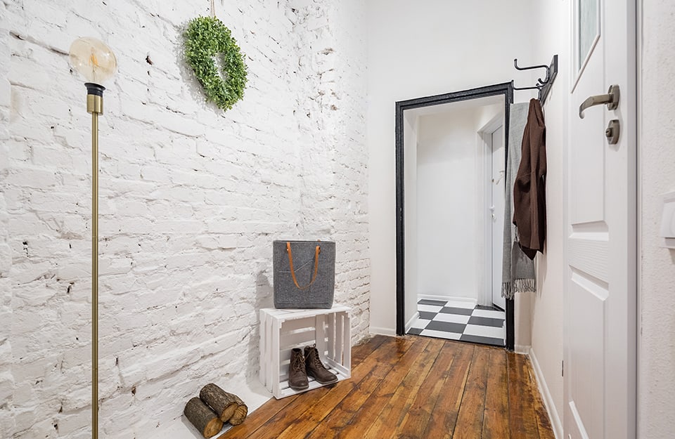 Ingresso di un abitazione. Le pareti sono in mattoni a vista, dipinti di bianco. A terra c'è un parquet rustico. Sul pavimento ci sono della scarpe e una cassetta da frutta dipinta di bianco. Una porta conduce a una stanza con pavimento a scacchiera bianco e nero