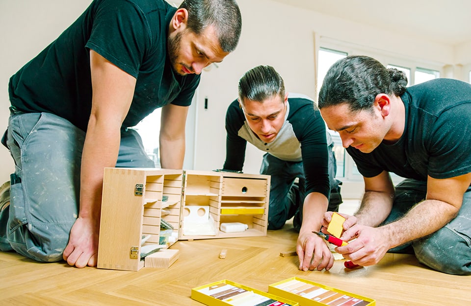 Tre uomini riparano le fessurazioni dei listoni di un parquet usando degli stick a cera