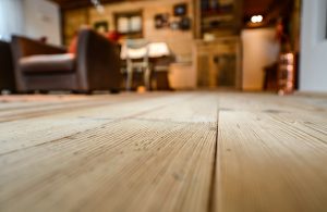 Dettaglio di un parquet rustico. Sfocato sullo sfondo si vede un salotto caldo e confortevole