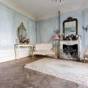 Ambiente shabby chic con arredi d'epoca riccamente decorati, pareti bianco e azzurro pastello, un grande tappeto davanti al caminetto e parquet rustico scuro