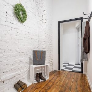 Ingresso di un abitazione. Le pareti sono in mattoni a vista, dipinti di bianco. A terra c'è un parquet rustico. Sul pavimento ci sono della scarpe e una cassetta da frutta dipinta di bianco. Una porta conduce a una stanza con pavimento a scacchiera bianco e nero