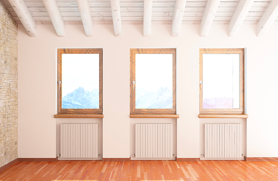 Mansarda di montagna con pavimento in parquet. Si vedono tre finestre affacciate sul panorama, con sotto tre termosifoni. Il soffitto è con travi a vista bianche e c'è una parete in pietra