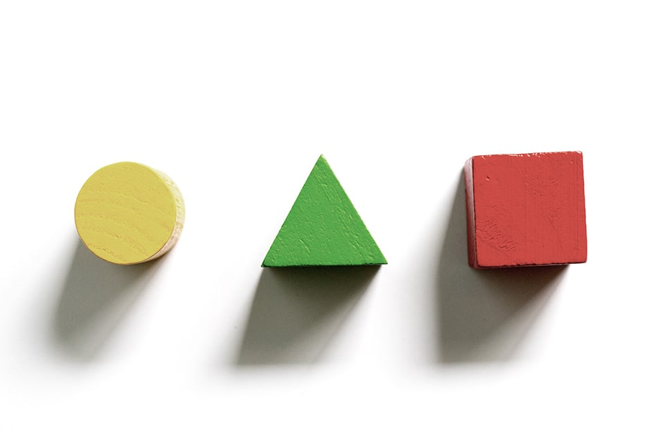 Tre forme geometriche tridimensionali in legno su fondo bianco: un cerchio giallo, un triangolo verde e un cubo rosso