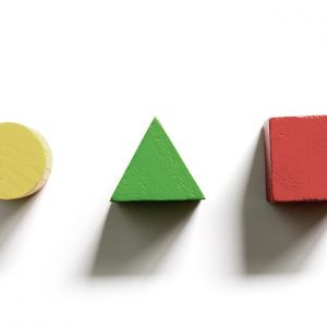 Tre forme geometriche tridimensionali in legno su fondo bianco: un cerchio giallo, un triangolo verde e un cubo rosso