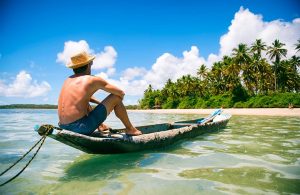 Uomo a torso nudo seduto rilassato su una piroga artigianale davanti a un'isola tropicale
