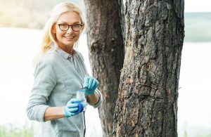 Una scienziata sorridente con gli occhiali mette un campione con una pinzetta in un barattolo, accanto a degli alberi in un bosco