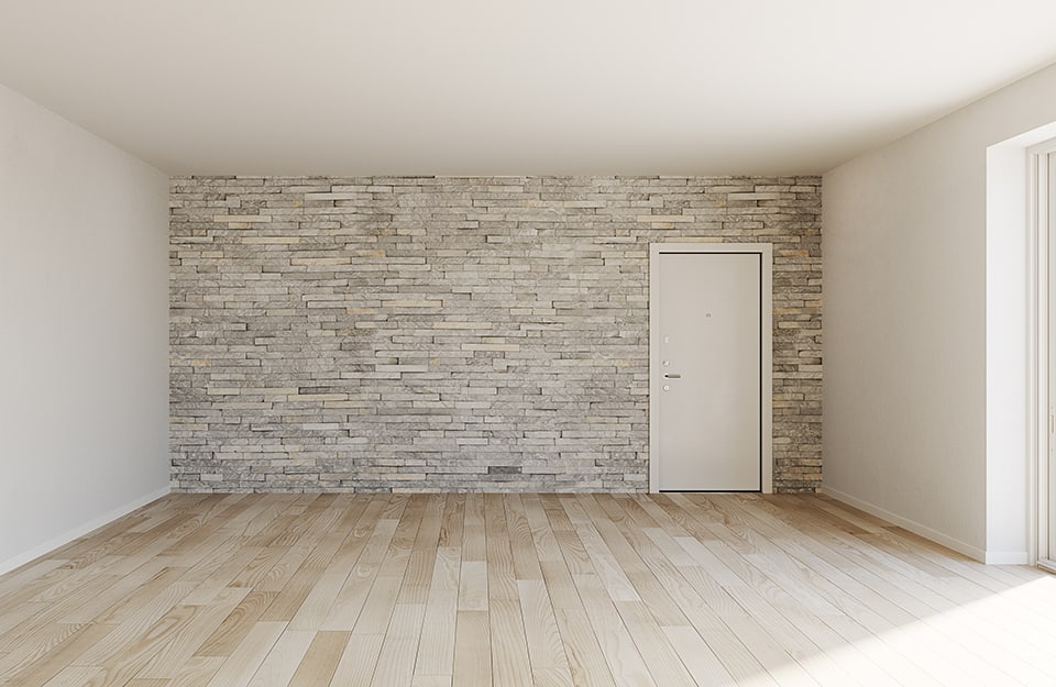 Stanza vuota con finestra da cui entra la luce, pareti e soffitto bianco e una parete con pietra a vista con porta bianca. Il parquet è in rovere chiaro