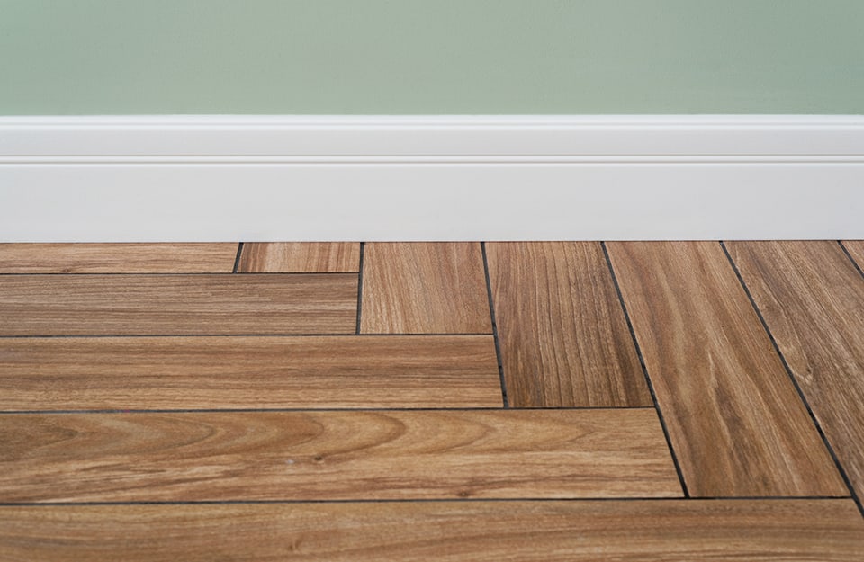 Dettaglio di un battiscopa bianco installato tra un muro verniciato di verde e un parquet a spina di pesce