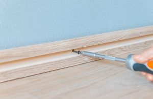 Un cacciavite che avvita un battiscopa alla parete, su un parquet chiaro