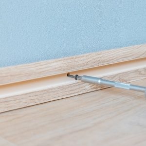 Un cacciavite che avvita un battiscopa alla parete, su un parquet chiaro