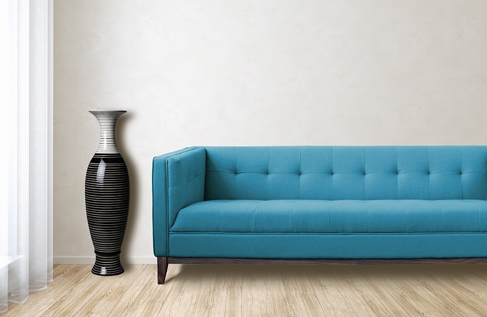 Salotto minimale con parquet in betulla, una finestra, un sofà trapuntato azzurro e un grande vaso nero e grigio dalle linee sinuose