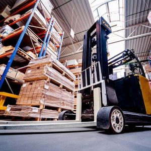 Un carrello elevatore visto dal basso, nel magazzino di un'industria di parquet piena di scaffali e pallet con assi di legno