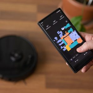 Robot aspirapolvere nero su un parquet, mentre una persona controlla l'apposita app sul suo smartphone