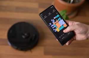Robot aspirapolvere nero su un parquet, mentre una persona controlla l'apposita app sul suo smartphone
