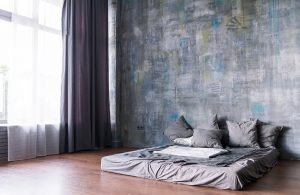 Luminosa stanza da letto iper-essenziale, con grande finestra, pareti in cemento grezzo, parquet, e materasso matrimoniale poggiato direttamente sul pavimento