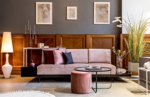 Salotto in stile vintage modernista con parete grigia, sofà rosa, sgabello rosa in tessuto, tavolino da caffè circolare in vetro e metallo nero, diverse piante, tre stampe astratte alle pareti abbinate tra loro e lampada da terra