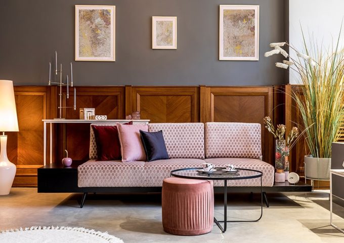 Salotto in stile vintage modernista con parete grigia, sofà rosa, sgabello rosa in tessuto, tavolino da caffè circolare in vetro e metallo nero, diverse piante, tre stampe astratte alle pareti abbinate tra loro e lampada da terra