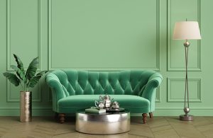 Angolo di un salotto in stile vintage con boiserie a cornici verde pastello, sofà trapuntato verde, tavolini e accessori metallizzati, lampada da terra e pianta in vaso
