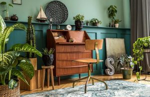 Area di un salotto con parquet a spina di pesce e armadio-scrittoio e sedia, boiserie in legno a mezza altezza di colore verde, su parete di un altro verde più chiaro. La stanza è molto carica di oggetti e piante e la boiserie funge anche da mensola per soprammobili