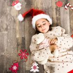 Una bambina con cappello di Babbo Natale e orsacchiotto tra le braccia, fotografata dall'alto sdraiata su un parquet con attorno delle decorazioni natalizie