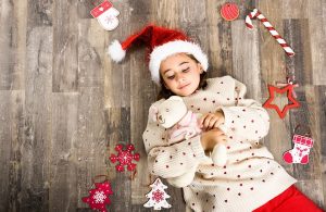 Una bambina con cappello di Babbo Natale e orsacchiotto tra le braccia, fotografata dall'alto sdraiata su un parquet con attorno delle decorazioni natalizie
