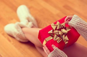 Una donna sta scartando un pacchetto regalo rosso con nastro d'oro. Indossa delle babucce pelose ed è sopra un parquet