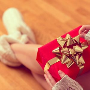 Una donna sta scartando un pacchetto regalo rosso con nastro d'oro. Indossa delle babucce pelose ed è sopra un parquet