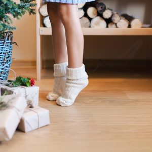 Una bambina in calzini di cui si vedono solo le gambe sta in punta dei piedi davanti accanto a un albero di Natale con sotto dei pacchi. Sullo sfondo c'è della legna per camino