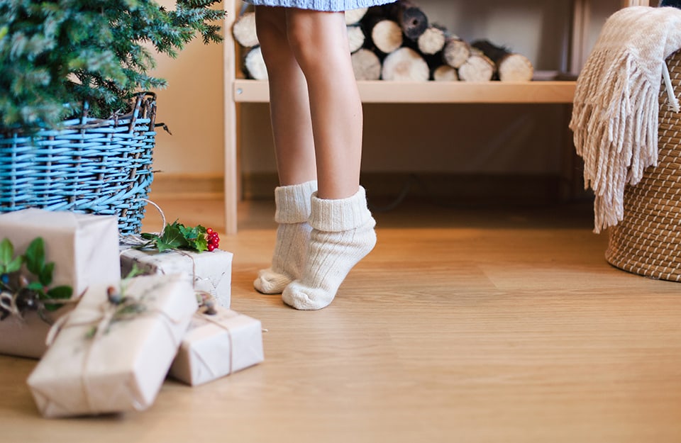 Una bambina in calzini di cui si vedono solo le gambe sta in punta dei piedi davanti accanto a un albero di Natale con sotto dei pacchi. Sullo sfondo c'è della legna per camino