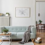 Salotto in stile moderno scandinavo con sofà celeste, tavolino da caffè in metallo bianco e legno, sgabello in legno con sopra una pianta, radiatore bianco in metallo con sopra una mensola e degli oggetti, una mappa incorniciata e appesa alla parete bianca e pavimento a parquet a spina di pesce. Oltre una porta si intravede una sala da pranzo